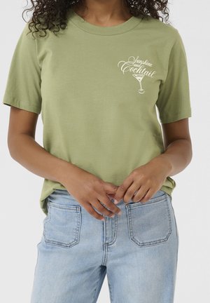 Mujer con camiseta verde claro con "Sunshine and Cocktail" y un gráfico de copa de martini, combinada con vaqueros vaqueros azul claro.