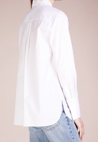 Mujer vistiendo una camisa blanca de manga larga holgada y jeans azul claro, vista desde atrás contra un fondo beige claro.