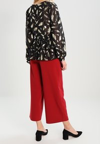 Blusa nera trasparente con stampa di foglie dorate, vita peplum; abbinata a pantaloni larghi rossi e scarpe col tacco nere.