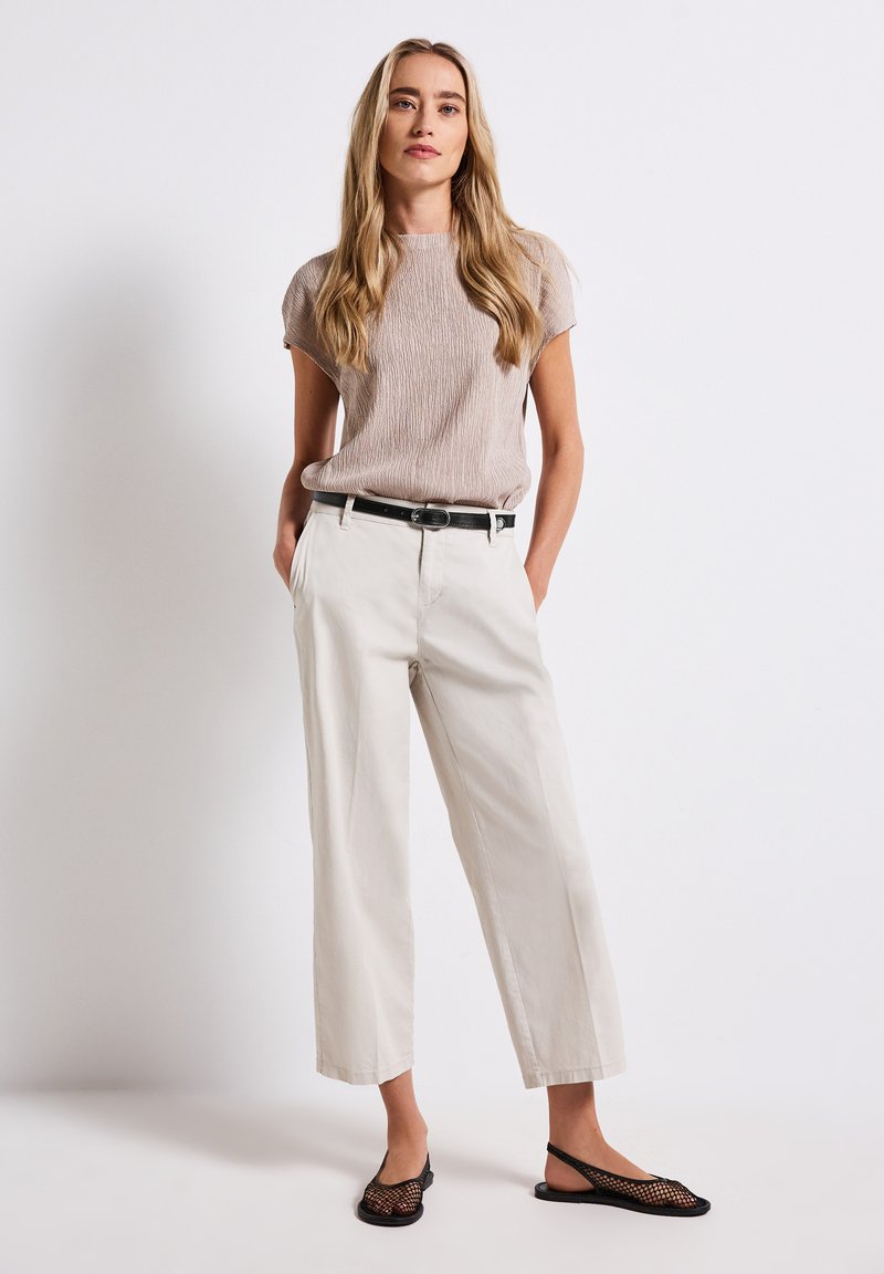 Beige tekstuuri top, valged laiad püksid, must vöö ja must võrkkangas slingback kingad. Mudel seisab käed taskutes neutraalse tausta ees.