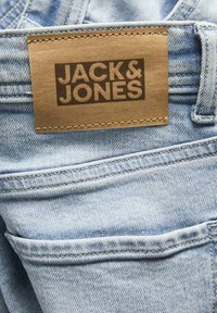 Jeans in denim azzurro chiaro con etichetta in pelle marrone con la scritta "JACK & JONES" in rilievo. Include tasche posteriori e dettagli di cucitura visibili.