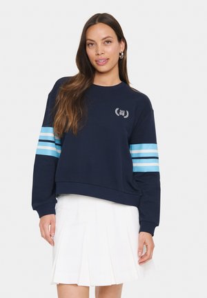Felpa navy con maniche a righe blu chiaro, scollo rotondo e logo ricamato. Indossata con una gonna bianca a pieghe.