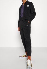 Chaqueta de pana negra con un parche de logo blanco, pantalones negros ajustados a juego y zapatillas coloridas con estampado. El conjunto tiene un acabado texturizado.