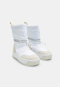 Witte gewatteerde laarzen met een hoge, geknoopte schacht, voorzien van een grijze stoffen accent en een beige suede neuskap, geplaatst op een witte rubberen zool.