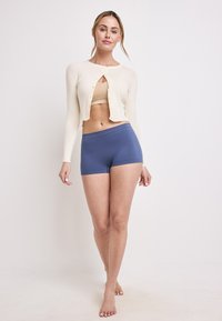 Ljus creamfärgad ribbad cardigan med rund halsringning och knappstängning, i kombination med blå shorts. Slät textur, avslappnad design, mid-crop längd.