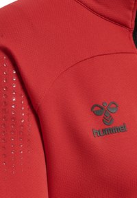 Rød præstationsjakke med struktureret stof og sort Hummel-logo. Har blanke prikdetaljer på den ene skulder for ekstra designaccent.