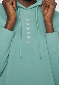 Händer justerar dragskoarna på en teal hoodie med vertikala silver Emporio Armani EA7-logotyper på bröstet.