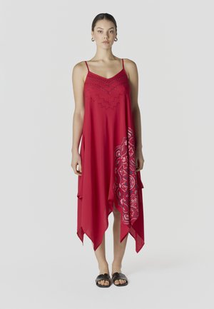 Mujer joven de pie con un vestido rojo sin mangas y falda asimétrica con estampado, y sandalias negras, sobre un fondo liso.