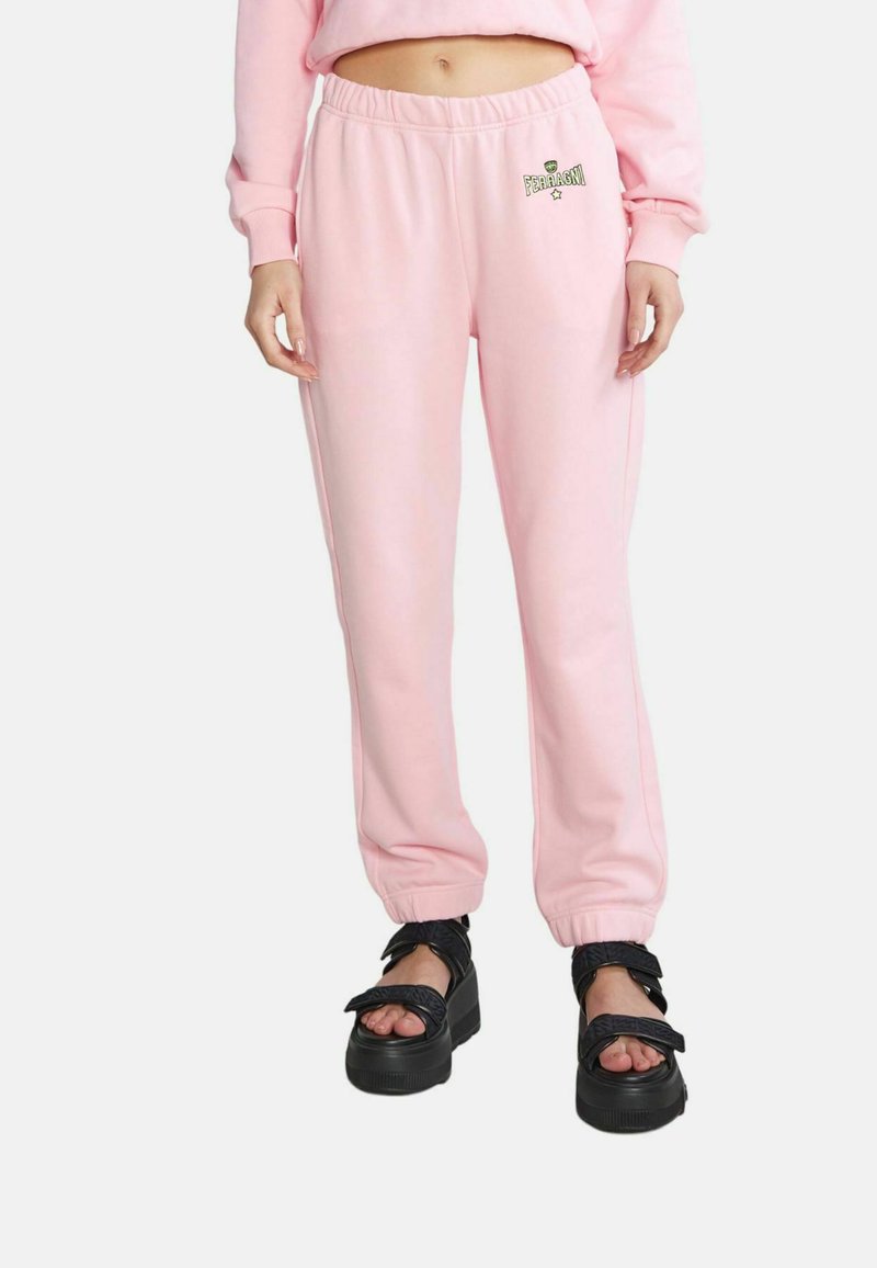 Rosa Jogginghose mit elastischem Bund und Bündchen am Knöchel, versehen mit einem kleinen grünen Logo. Kombiniert mit Plateausandalen und einem oversized Oberteil.
