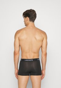Black cotton boxer shorts with a solid design. Features a wide elastic waistband with "LACOSTE" logo. Smooth texture, fitted shape.