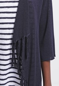 Tmavě modrý přehoz s ohraničenými okraji a měkkou pletenou texturou. Pod ním bílá košile s tmavě modrými vodorovnými pruhy.