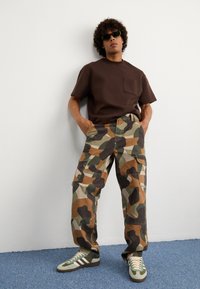 Ruskea lyhythihainen t-paita, jossa on tasku, yhdistettynä monivärisiin kamouflage cargo-housuihin ja vihreäviiruisiin lenkkareihin valkoisilla yksityiskohdilla.