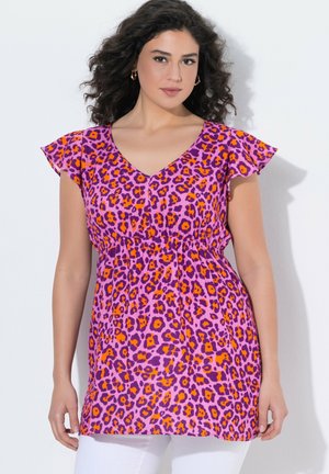 Kobieta o kręconych włosach nosi fioletową i pomarańczową tunikę w kwiatowy wzór z luźnymi rękawami oraz białe spodnie, stoi na białym tle.