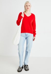 Pull rouge en tricot à col en V, avec des manches longues, associé à un jean droit bleu clair. Sac à épaule texturé blanc et chaussures noires à imprimé zèbre.