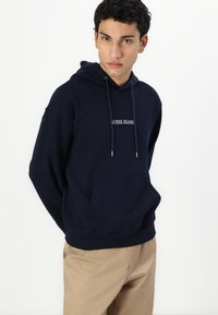 Tengerészkék kapucnis pulóver pamut keverékből, elöl kenguruzsebbel, húzózsinórral ellátott kapucnival és fehér "GUESS JEANS" logóval.
