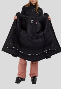 Sort isoleret jakke med en glat tekstur og elastiske ærmer, der har flere indvendige lommer og branding. Bæres over lyserøde skibukser.