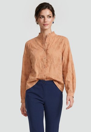 Frau trägt eine pfirsichfarbene Bluse mit Knöpfen und floraler Stickerei sowie dunkelblaue Hose, steht vor einem einfarbigen hellen Hintergrund.