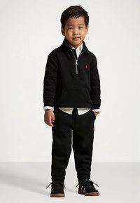 Young boy standing wearing a black half-zip sweatshirt, white shirt underneath, black pants, and black sneakers with brown soles.