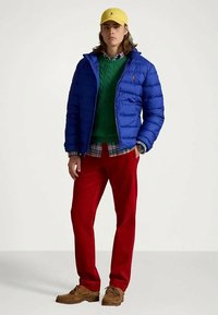 Jovem vestido com um boné amarelo, uma jaqueta puffer azul, um suéter verde, uma camisa à quadrados, calças vermelhas e sapatos castanhos, em pé com a mão no bolso.