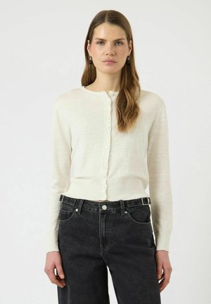 Mujer con cabello largo y castaño que lleva un cárdigan blanco abotonado y jeans negros de talle alto, posando frente a un fondo claro y liso.