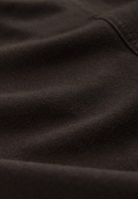 Gros plan d’un tissu doux brun foncé avec une texture tissée visible et de subtils plis soulignant la qualité du matériau.