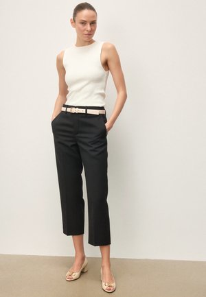 Mujer con el cabello recogido, vistiendo una camiseta blanca sin mangas, pantalones negros recortados con cinturón blanco y zapatos beige abiertos con tacón bajo, de pie.