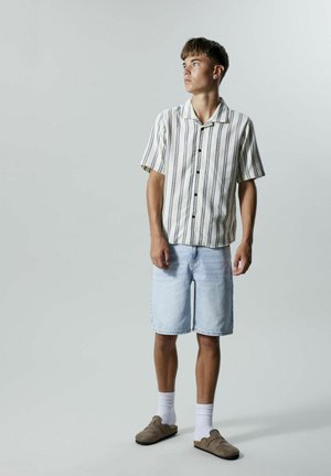 Šviesiai mėlynos džinsinės šortai ir trumpomis rankovėmis dryžuota palaidinė smėlio ir jūros gylio spalvomis, derinamos su rudais šlepetėmis ir baltais kojiniais.