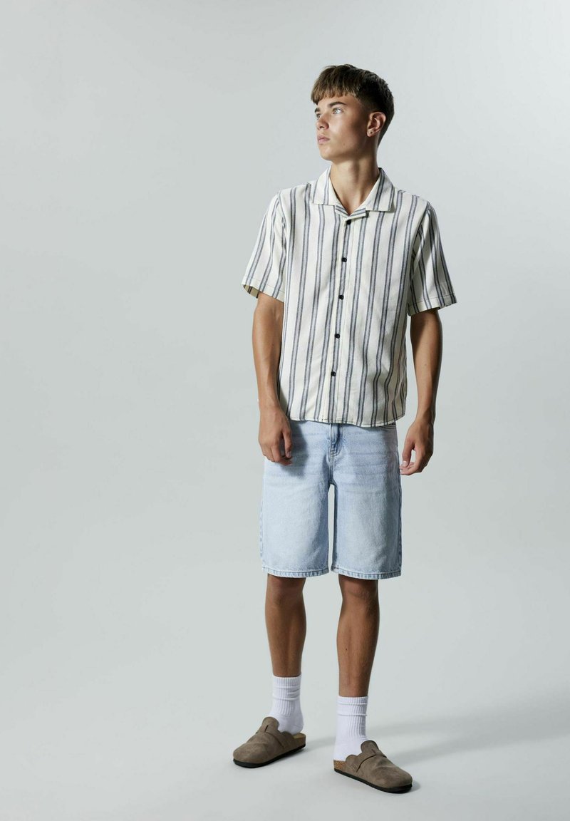 Kortermet, stripete skjorte i beige og marineblå, sammen med lyseblå denimshorts og brune slip-on-sko, med hvite sokker.