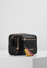 Černá kožená taška se zlatými detaily, dvěma zipovými přihrádkami a barevnými třásněmi v růžové a žluté barvě na táhlech zipu.