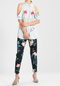 Camicia azzurro chiaro con spalle scoperte e ricami floreali, abbinata a pantaloni neri con un motivo floreale colorato. Tacchi argento.