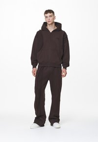 Sudadera con cremallera de color marrón, con una gran capucha y bolsillos frontales, combinada con pantalones a juego que cuentan con costuras laterales; textura suave y corte relajado.