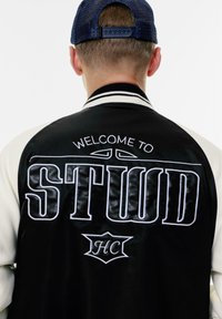 Veste varsity en noir et blanc avec le texte brodé "WELCOME TO STUUD" et un détail de logo au dos ; elle possède un col côtelé et un tissu texturé.