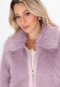 Giovane donna con capelli biondi che indossa orecchini a cerchio dorati e una giacca con zip fluffy color lavanda sopra un top in pizzo bianco.