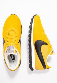 Gula Nike-skor med en svart Swoosh, i mocka och mesh-material, texturerad svart sula och platta snören. Märkbara vita detaljer på tungan.