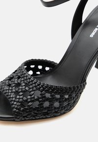 Μαύρο σανδάλι με ψηλό τακούνι και πλεκτό δερμάτινο λουράκι πάνω από τα δάχτυλα, λείο πέλμα, μερικώς ορατό λουράκι στον αστράγαλο.