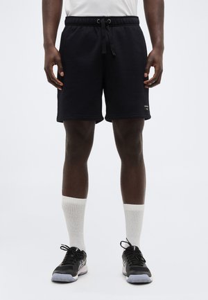 Mees, kes kannab musta paelaga lühikesi pükse, valgeid keskmise sääre pikkusega sokke ja musti spordijalatsid, seisab ühevärvse tausta ees.