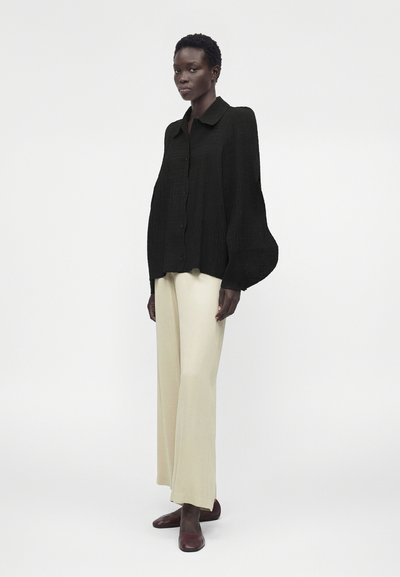 Γυναίκα με μαύρο πουκάμισο με μακριά μανίκια και φουσκωτά μανίκια, κρεμ παντελόνι με φαρδιά πόδια και σκούρα καφέ επίπεδα παπούτσια, στέκεται σε λευκό φόντο.