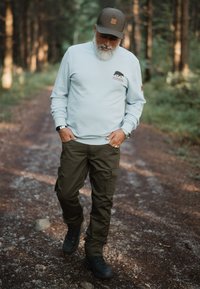 Sudadera azul claro con el logo "Forsberg", combinada con pantalones cargo verde oliva y botas negras, llevada por una persona que camina por un camino de grava.