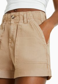 Vaaleat beiget lyhyet housut, jotka on valmistettu kevyestä kankaasta. Niissä on etutaskut, nappikiinnitys ja käännetyt lahkeet, joissa on näkyviä ompelutyynyjä.