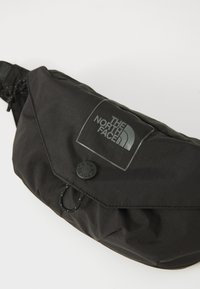 Schwarze Nylon-Hüfttasche mit Klappenverschluss, ausgestattet mit einem strukturierten Logo-Patch und einer Kordelzugdetail. Glatte Oberfläche, unaufgeregtes Design.
