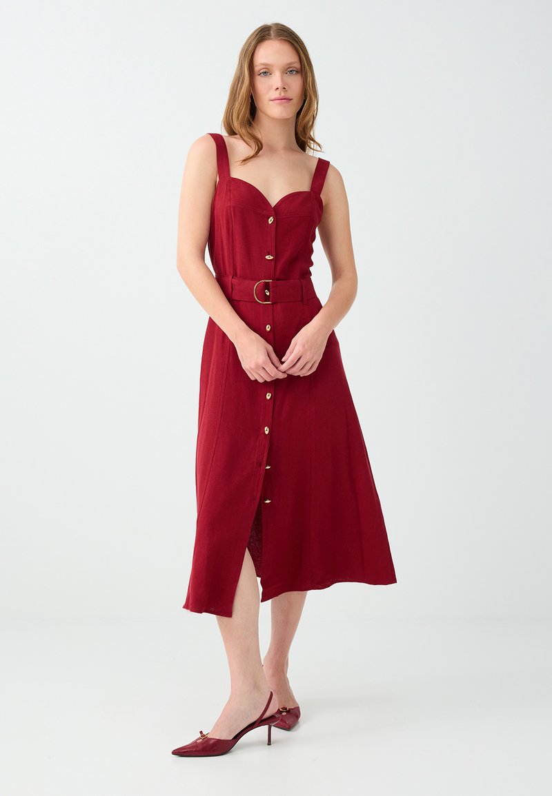 Femme portant une robe midi rouge sans manches avec des boutons sur le devant et une ceinture, associée à des talons slingback rouges assortis.