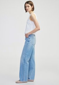 Jeans de mezclilla anchos de color azul claro con un patrón de manchas blancas, con una cintura alta, bolsillos laterales y un corte relajado. Combinados con una blusa sin mangas blanca.