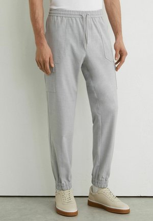 Jasnoszare spodnie joggery cargo z elastycznym pasem i ściągaczami, noszone z beżowymi sneakersami i białymi skarpetkami na stojącej osobie.