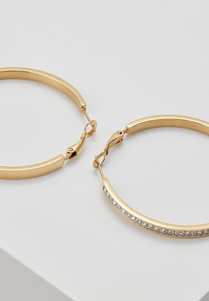 Boucles d’oreilles créoles en or, dont une présente une rangée de petites pierres précieuses claires le long du bord extérieur, exposées sur une surface blanche.