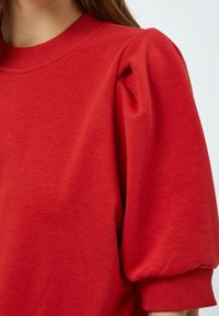 Žena v červeném svetru s vycpanými rukávy a žebrovaným kulatým výstřihem, zobrazená od ramene po horní část paže.