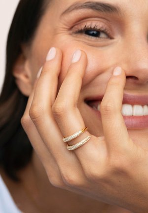 Anillos de tono dorado con cristales claros y brillantes, con un diseño liso y curvado. Los anillos están apilados juntos en una mano cerca de un rostro sonriente.