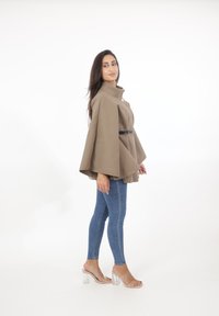 Brun, överdimensionerad cape-liknande topp med hög krage och frontknappdetalj, parat med blå skinny jeans och klara blockklacksandaler.