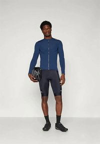 Ρούχο ποδηλασίας με εφαρμοστό ναυτικό μπλουζάκι μακρυμάνικο με φερμουάρ μπροστά, συνδυασμένο με μαύρες ποδηλατικές βερμούδες και μαύρα ποδηλατικά παπούτσια, κρατώντας ένα κράνος.