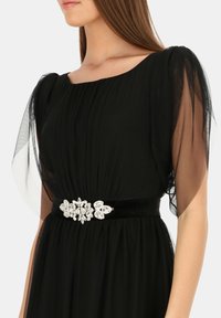 Czarna wieczorowa sukienka z przezroczystymi, bufkami, marszczonym gorsetem i aksamitnym paskiem ozdobionym kryształową aplikacją w kształcie kwiatu.