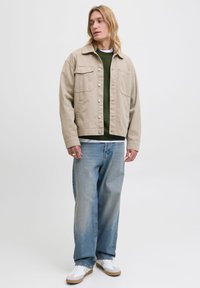 Ung person med axellångt blont hår som bär en beige jacka, grön tröja, ljusblå vida jeans och vita sneakers mot vit bakgrund.